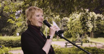 Film still from 'A portrait of Alex Post' (2024) by Jon Beaton. A non-binary person speaking into a microphone in a park, smiling.